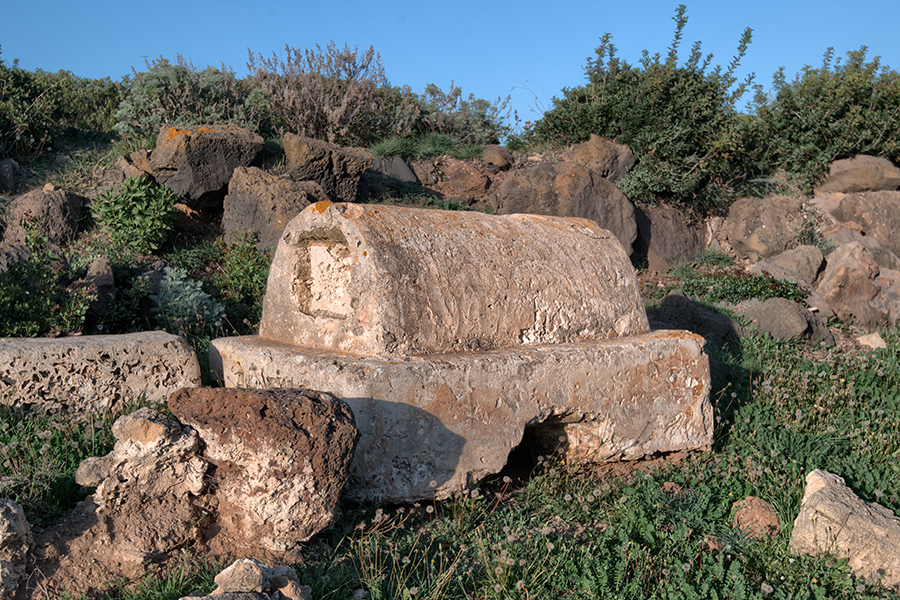 Ruins of an Imperial Roman necropolis in Tharros. A domed stone tomb surrounded by stone fragments on the grass.